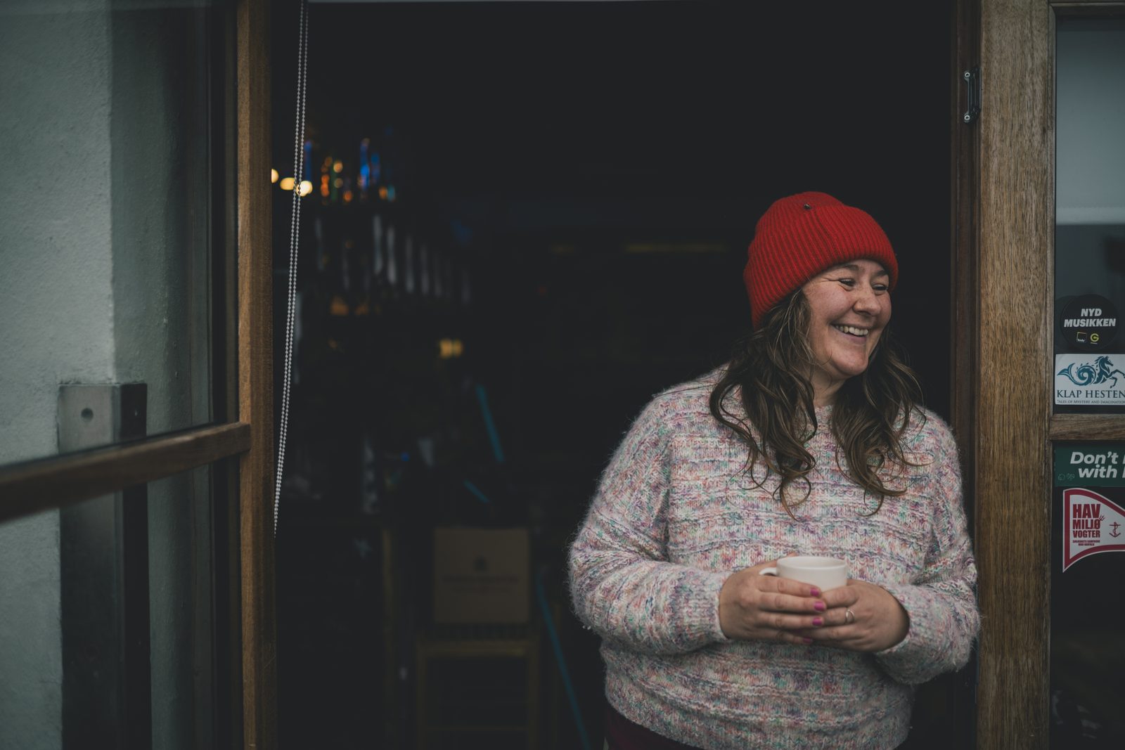 Woman in a red hat holding a mug, laughing in the doorway of a café in Klintholm Havn