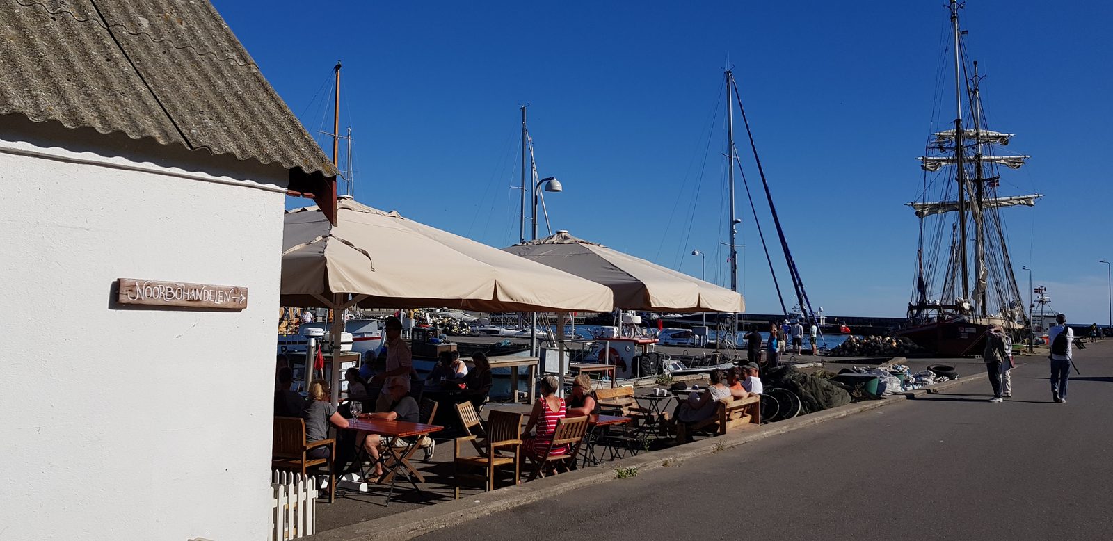Guests under parasols at Klintholm Havn harbour, boat masts behind
