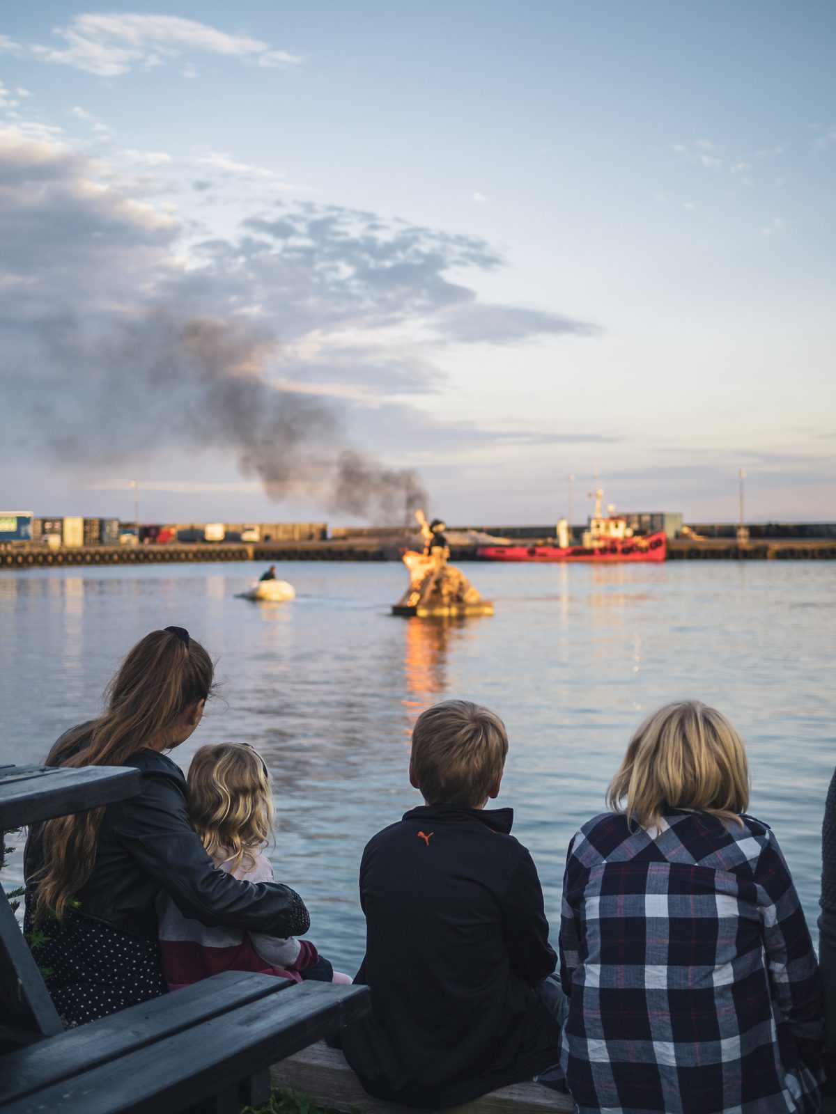 A family seen from behind sitting at the quay, watching a bonfire on a raft in the harbour basin
