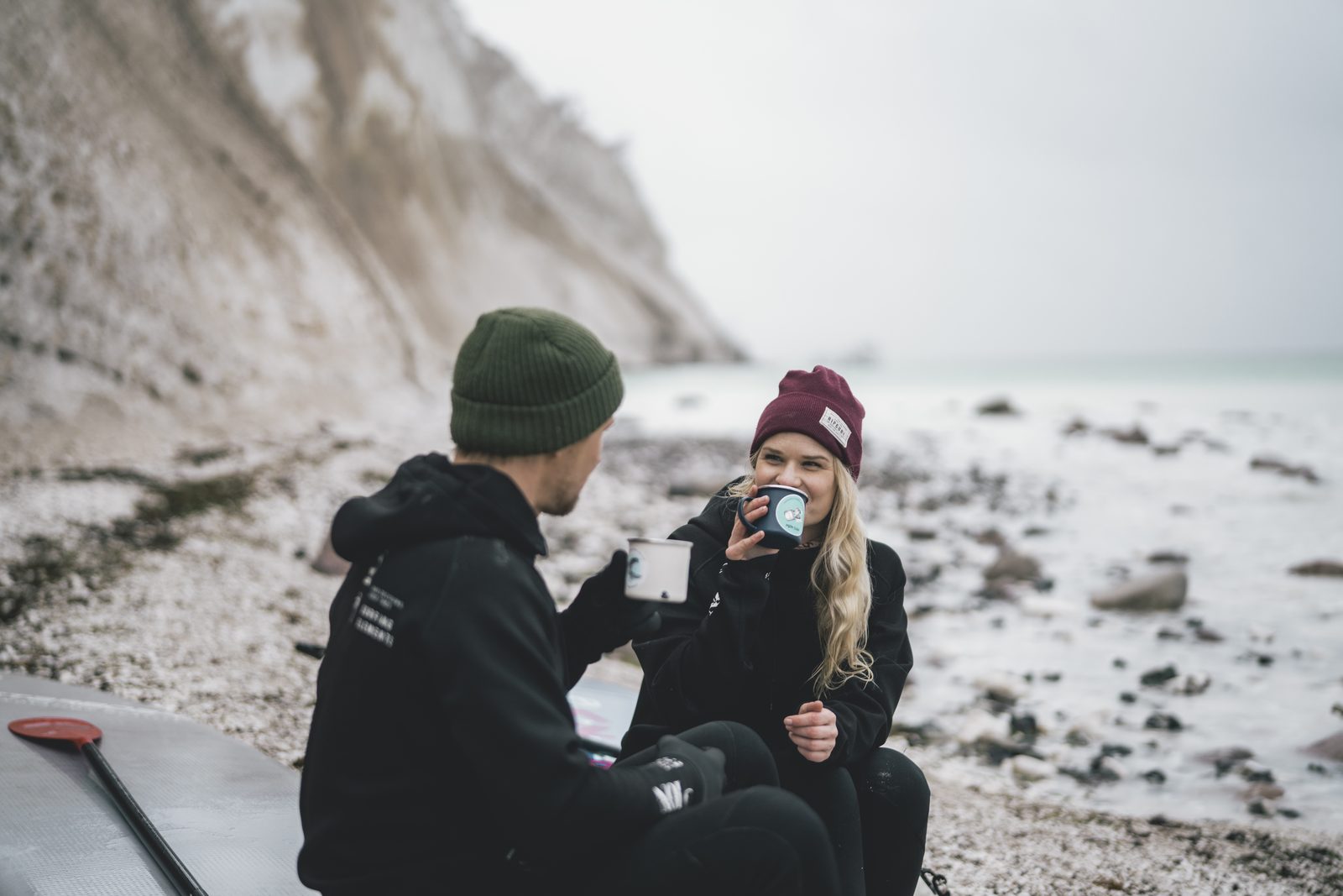 Two people in winter clothes drinking from enamel mugs on the beach in front of the chalk cliff
