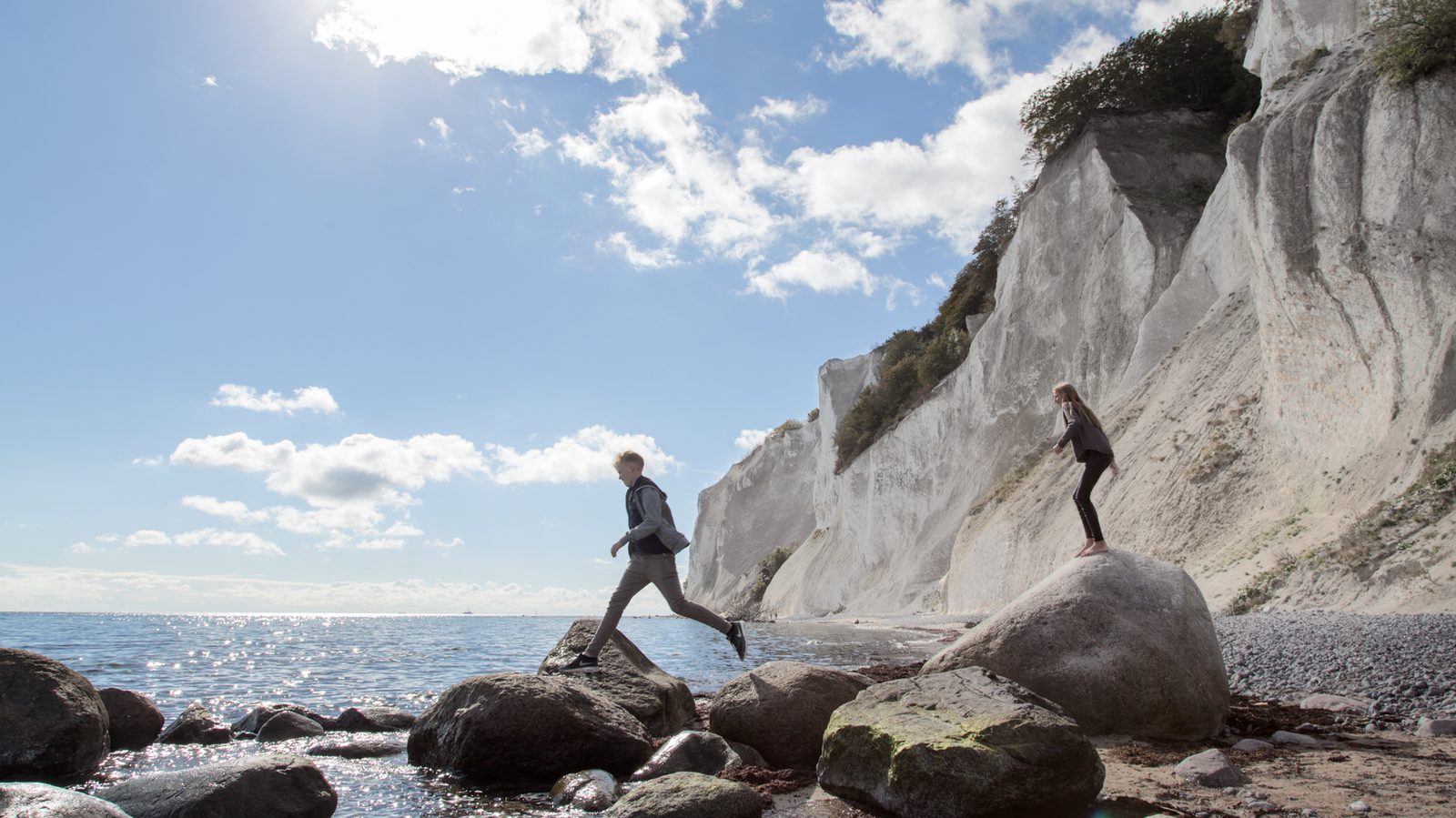 Two visitors hopping across boulders on the beach below the chalk cliffs of Møns Klint
