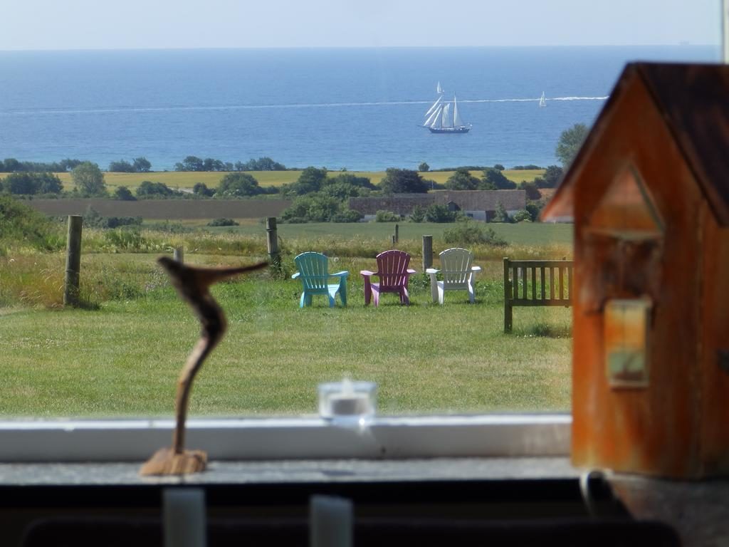 Idyllischer Ausblick von Bakkegaard Møns Klint mit Blick auf die Ostsee. Im Vordergrund eine rustikale Skulptur und ein Vogelhaus, dahinter eine Gruppe farbenfroher Gartenstühle auf einer Wiese. Im Hintergrund segelt ein Großsegler auf dem Meer