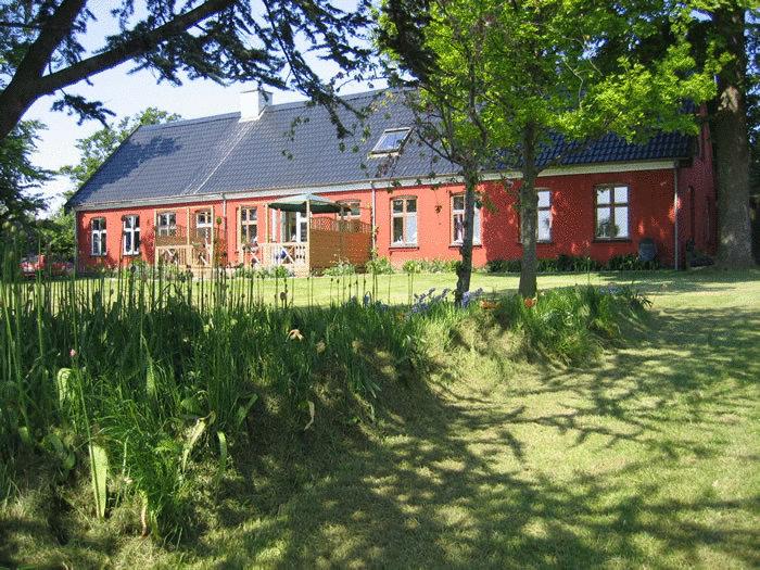 Blick auf das Bakkelund Bed & Breakfast auf der Insel Mön, ein einstöckiges, rotes Ziegelhaus mit blauem Dach, umgeben von einem gepflegten Garten mit Grasflächen und hohen Bäumen. Das ländliche Anwesen strahlt Ruhe und Gemütlichkeit aus.