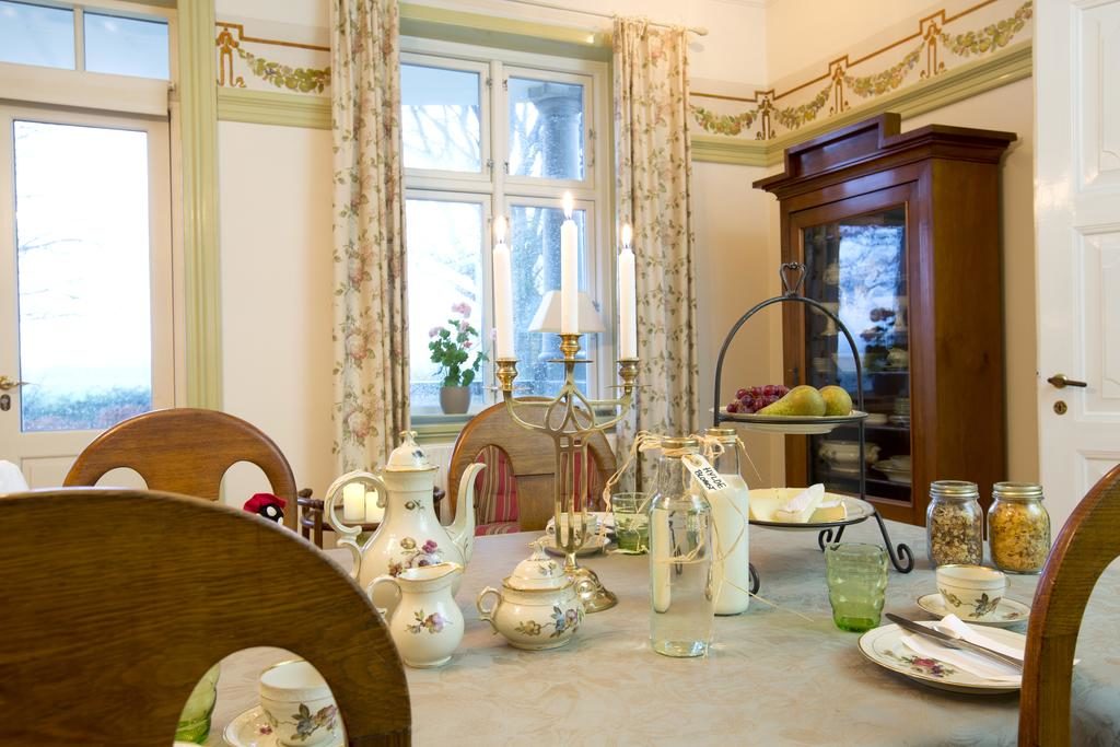 Elegant gedeckter Frühstückstisch im Tiendegaarden Møn, mit einer Auswahl an Porzellangeschirr und Silberbesteck. Der Raum ist hell und einladend, mit großen Fenstern, die den Blick auf den Garten freigeben. Klassisches Interieur und liebevoll arrangierte Dekorationen schaffen eine warme Atmosphäre.