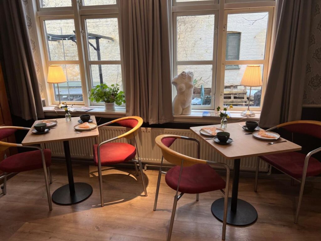 Stilvoll eingerichtetes Café im Apothekergaarden in Stege, mit mehreren kleinen Tischen, die für das Frühstück gedeckt sind. Jeder Tisch ist mit roten und hölzernen Stühlen ausgestattet, neben einem Fenster, durch das Tageslicht strömt. Eine Skulptur und Zimmerpflanzen verleihen dem Raum ein künstlerisches Flair.