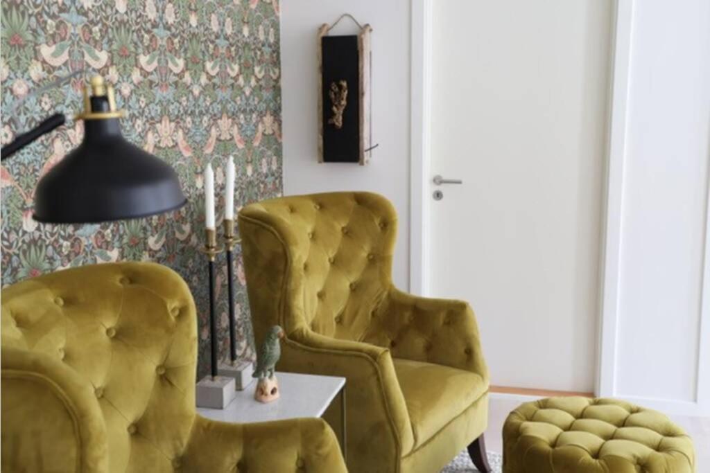 Elegantes Leseecke-Design im Lejlighed på Torvet in Stege mit zwei üppigen, samtigen, olivgrünen Sesseln vor einer mit floralem Muster tapezierten Wand. Ergänzt wird die Szene durch eine moderne schwarze Stehlampe und einen passenden Hocker. Dieser gemütliche Bereich lädt zum Entspannen und Lesen ein.