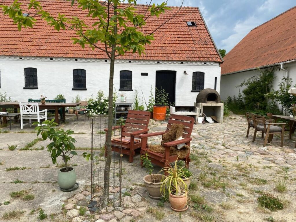 Rustikaler Innenhof des Camønøgården Bed & Breakfast, geprägt durch einen Kopfsteinpflasterhof umgeben von weißen Gebäuden mit dunklen Fensterläden. Im Hof sind mehrere Sitzgruppen verteilt, eine davon neben einem alten Steinofen, was eine gemütliche Atmosphäre für gesellige Abende bietet.
