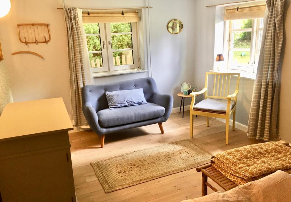 Gemütlicher Wohnbereich im Hvidegaard Bed & Breakfast, ausgestattet mit einem modernen grauen Sofa, einem gelben Stuhl und einem kleinen Holztisch. Der Raum ist hell, mit Holzböden und sanften Farben dekoriert und blickt auf einen grünen Garten durch große Fenster.